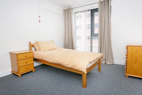 Simple bedroom with a single wooden bed, matching bedside table and chest of drawers. A window with floor‑length curtains lets in natural light, and the room is carpeted with neutral décor.