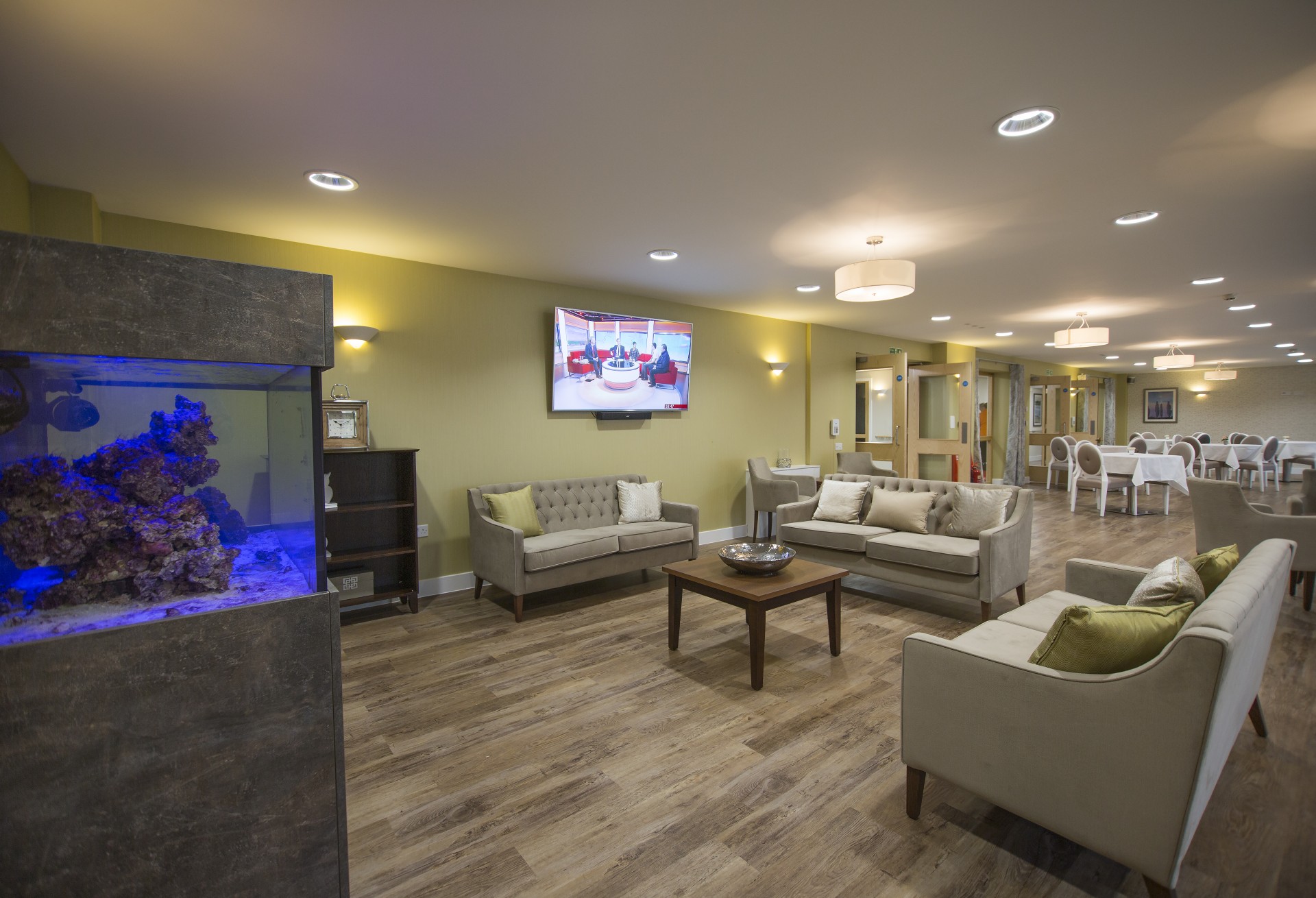 Communal lounge with sofas arranged around a wooden coffee table, set beside a large built‑in aquarium with blue lighting. A television is mounted on a green feature wall, with wooden flooring and doors leading to a dining area at the back.