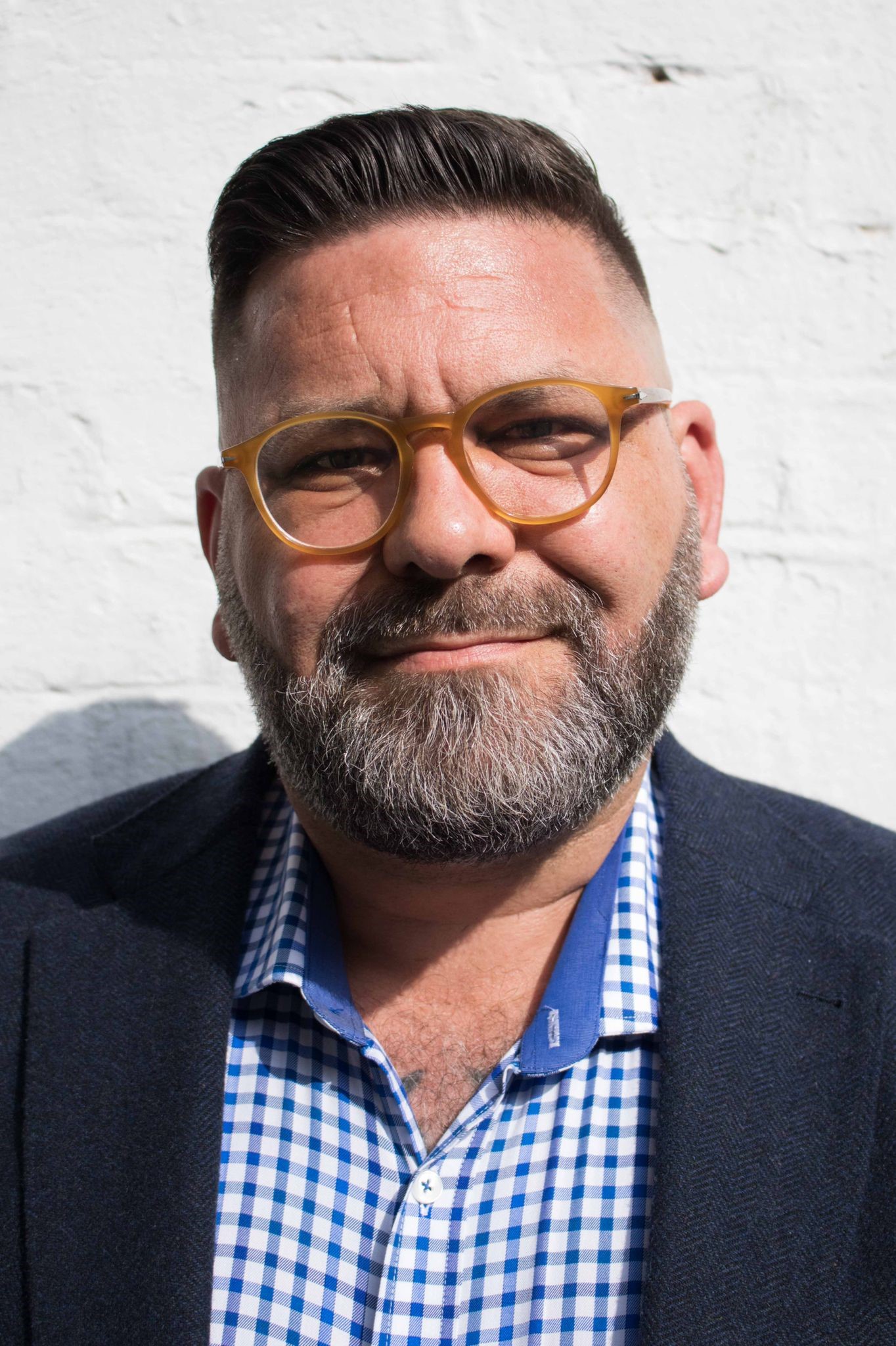 Portrait of a bearded man with short dark hair wearing amber‑coloured glasses, a checked shirt and a dark jacket. He stands in front of a white wall, smiling slightly at the camera.