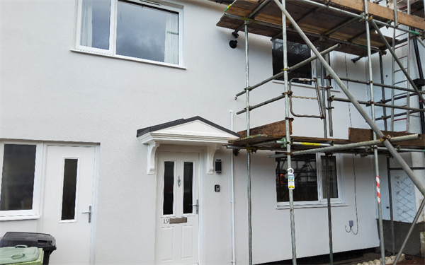 Exterior of a two‑storey house with a white front door and small porch canopy. Scaffolding is erected along the front of the building, indicating maintenance or repair work in progress, with windows visible on both floors.