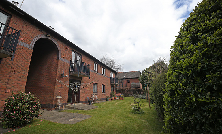 Exterior of a two‑storey red‑brick residential building with wooden‑framed windows and small balconies, viewed across a communal garden. A neatly kept lawn runs along the building, with patio seating, potted plants and a rotary clothes airer near the entrances. A tall hedge borders the right side of the garden, with additional residential buildings visible in the background under a cloudy sky.