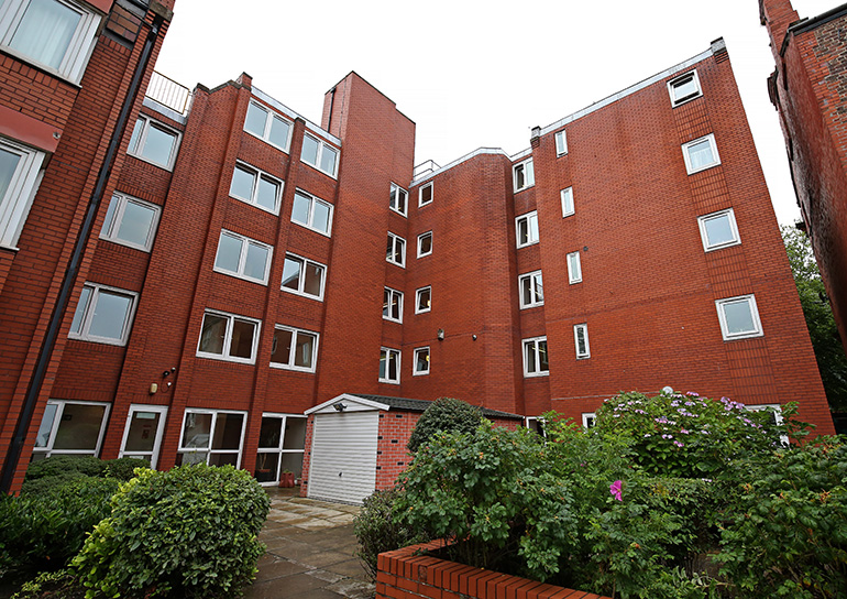 Exterior of a multi‑storey red‑brick residential building with white‑framed windows, arranged around a planted garden area. Low brick walls, shrubs and flowering plants line a paved path leading toward the entrance, with greenery in the foreground and neighbouring building sections visible under an overcast sky.