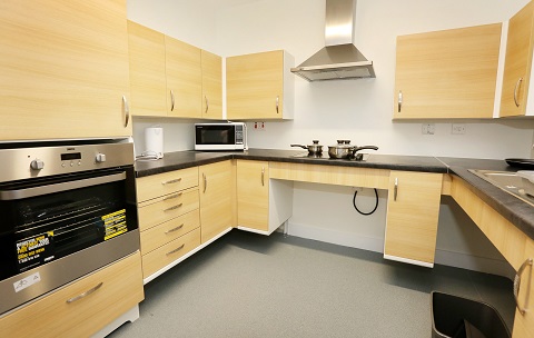 Modern fitted kitchen with light wood cabinets, grey worktops and integrated appliances including an oven, hob and extractor hood. A microwave and kettle sit on the worktop, with vinyl flooring throughout.