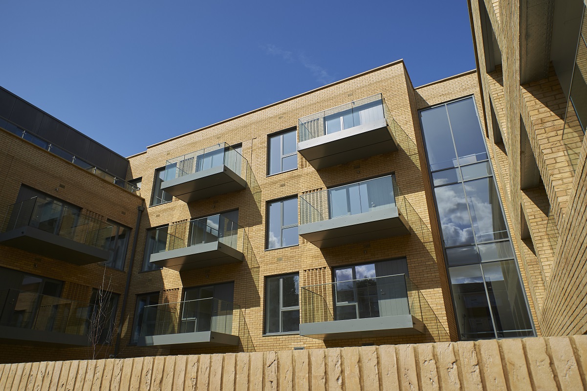 Exterior of a modern multi‑storey residential building with light‑coloured brick walls and glass‑fronted balconies. Large windows line the façade, with a clear blue sky above and a low brick wall in the foreground.