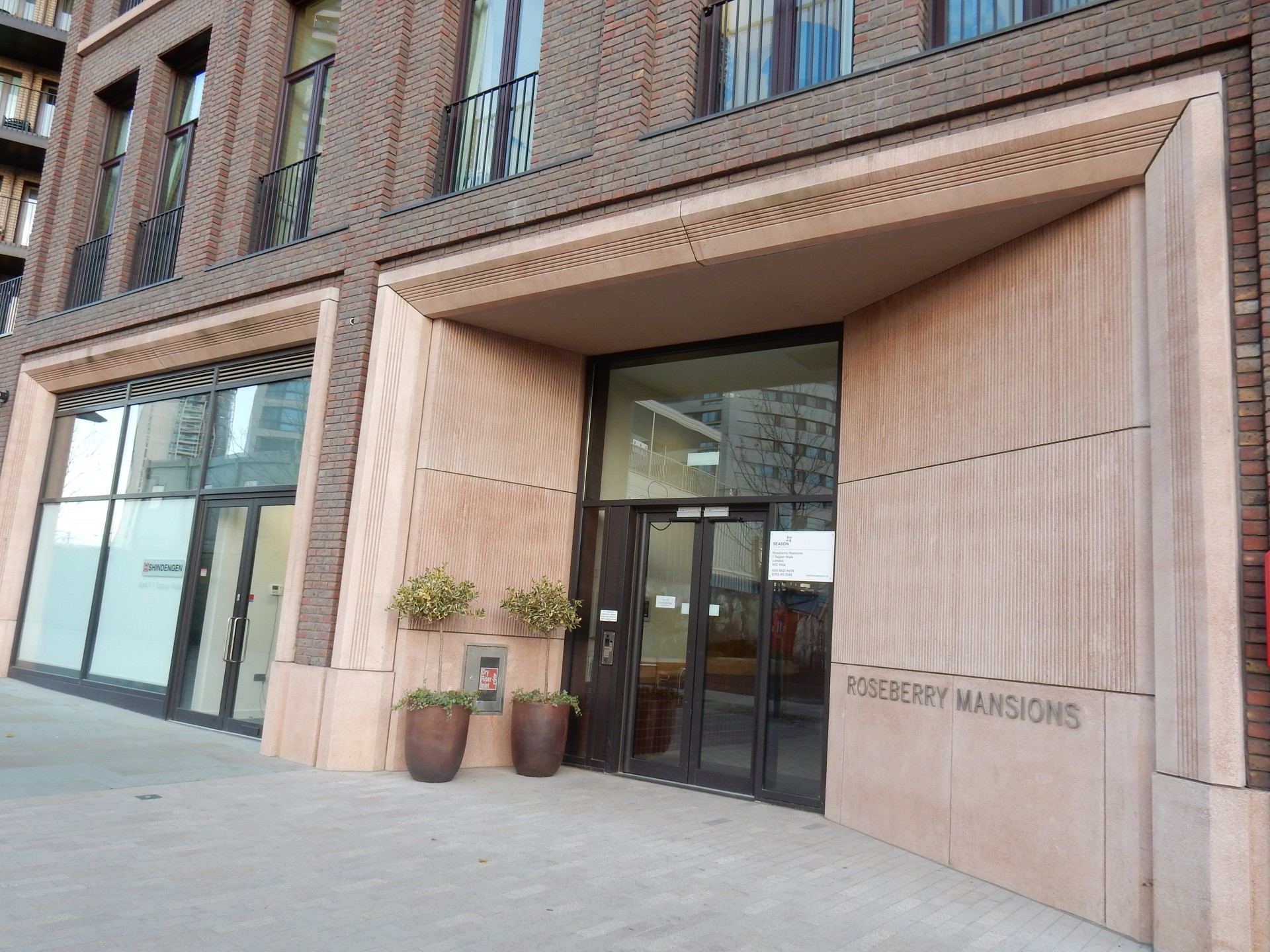 Entrance to a modern residential building with stone‑framed doorway and large glass doors. A sign on the wall reads “Roseberry Mansions,” with large potted plants positioned beside the entrance and brick façades with balconies rising above.