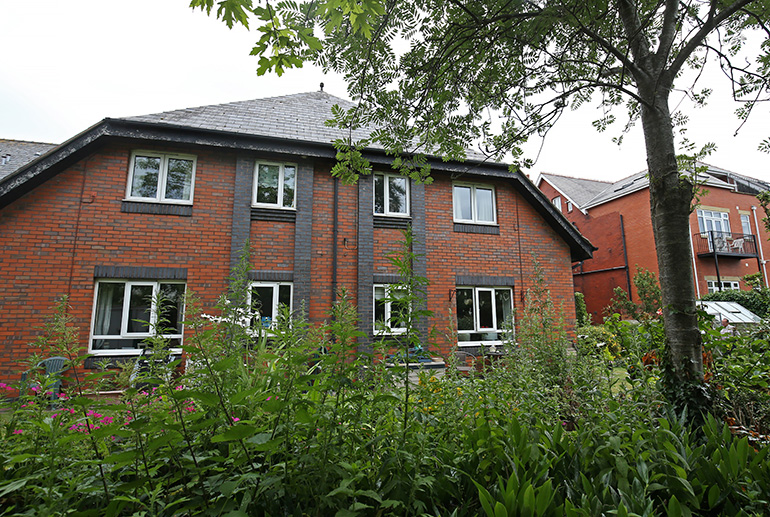 Exterior of a two‑storey red‑brick residential building with white‑framed windows, viewed through a landscaped garden with tall leafy plants and flowers in the foreground. Mature trees frame the scene, with additional residential buildings visible nearby under an overcast sky.