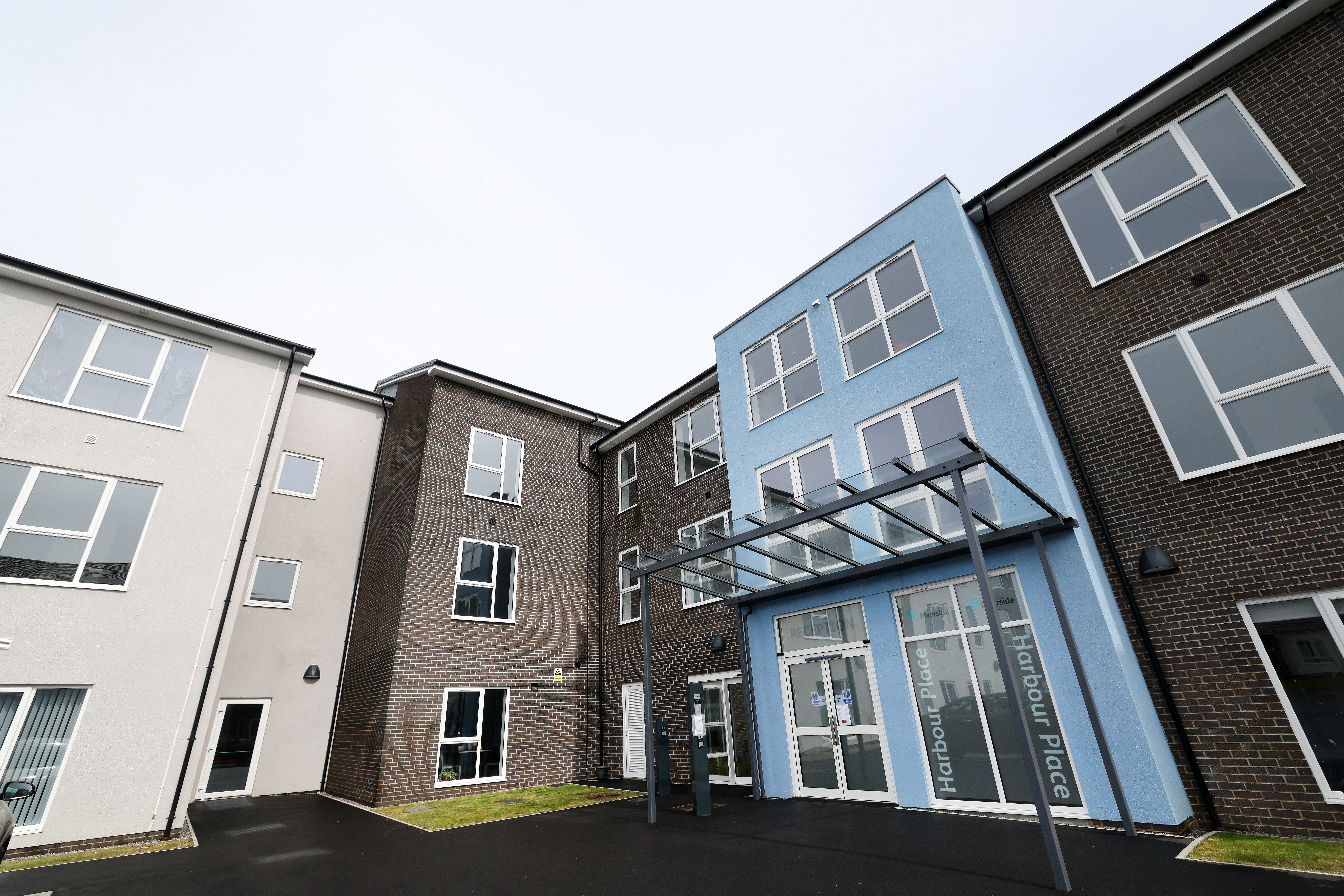 Exterior of a modern multi‑storey residential building with brick and light‑coloured cladding. A blue entrance section with a glass canopy marks the main doors, with white‑framed windows and paved access areas around the building.