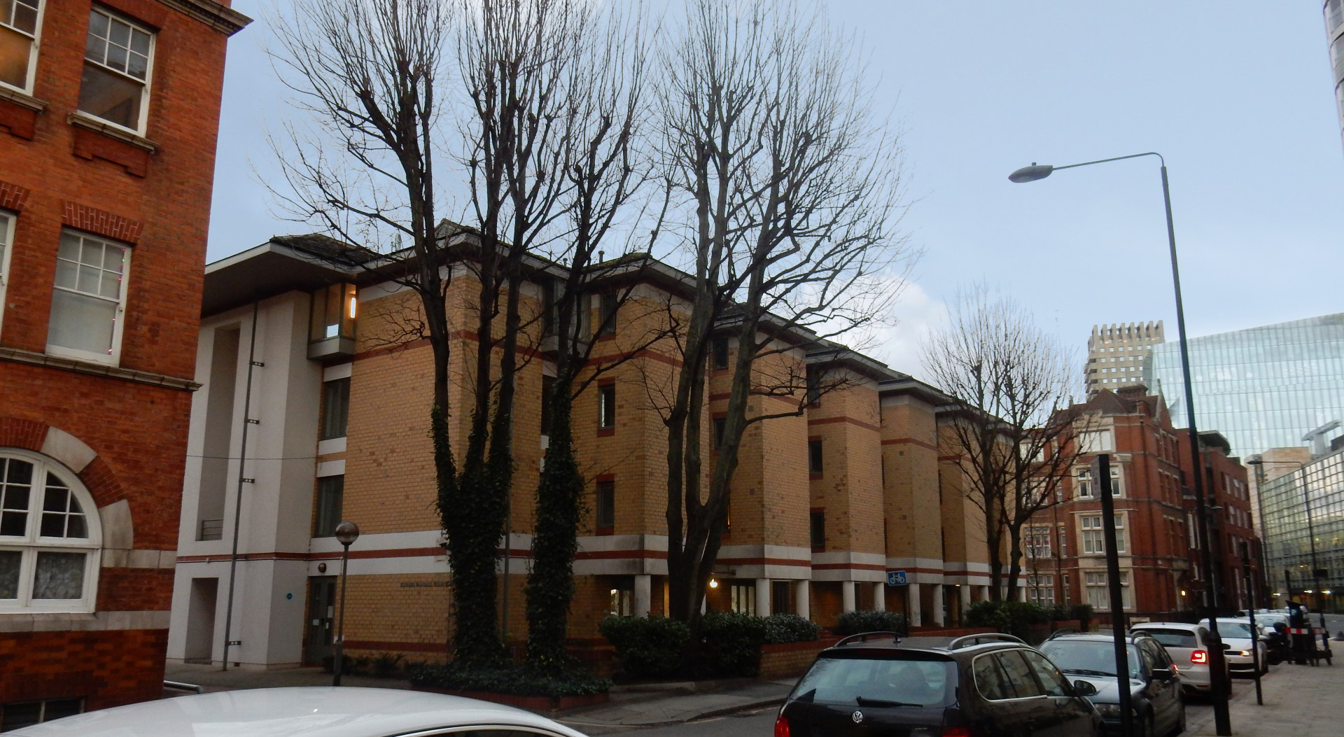 Street view of a residential building with light tan brick and white structural columns at ground level, creating a covered walkway. Leafless trees line the pavement in the foreground, with parked cars along the street and a mix of older red‑brick buildings and modern glass offices visible in the background under an overcast sky.
