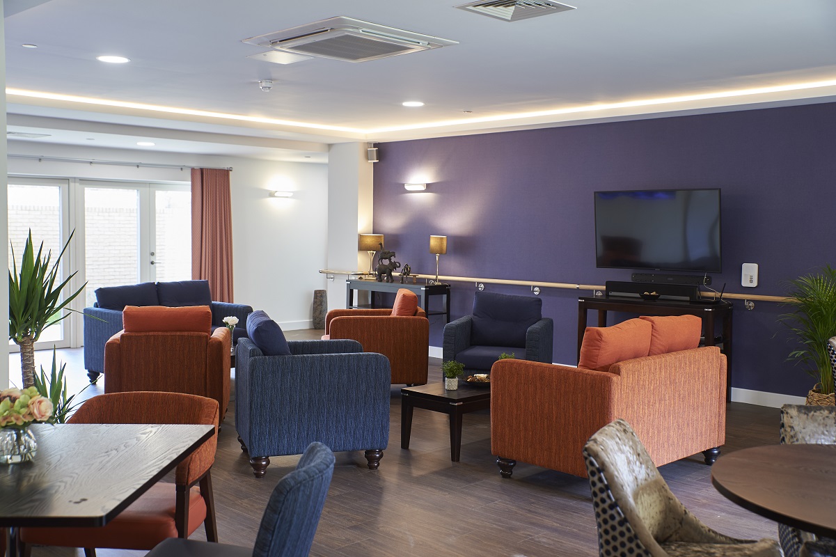Communal lounge with armchairs and sofas in orange and blue arranged around low coffee tables. A television is mounted on a purple feature wall, with soft lighting, wooden flooring and glass doors letting in natural light.