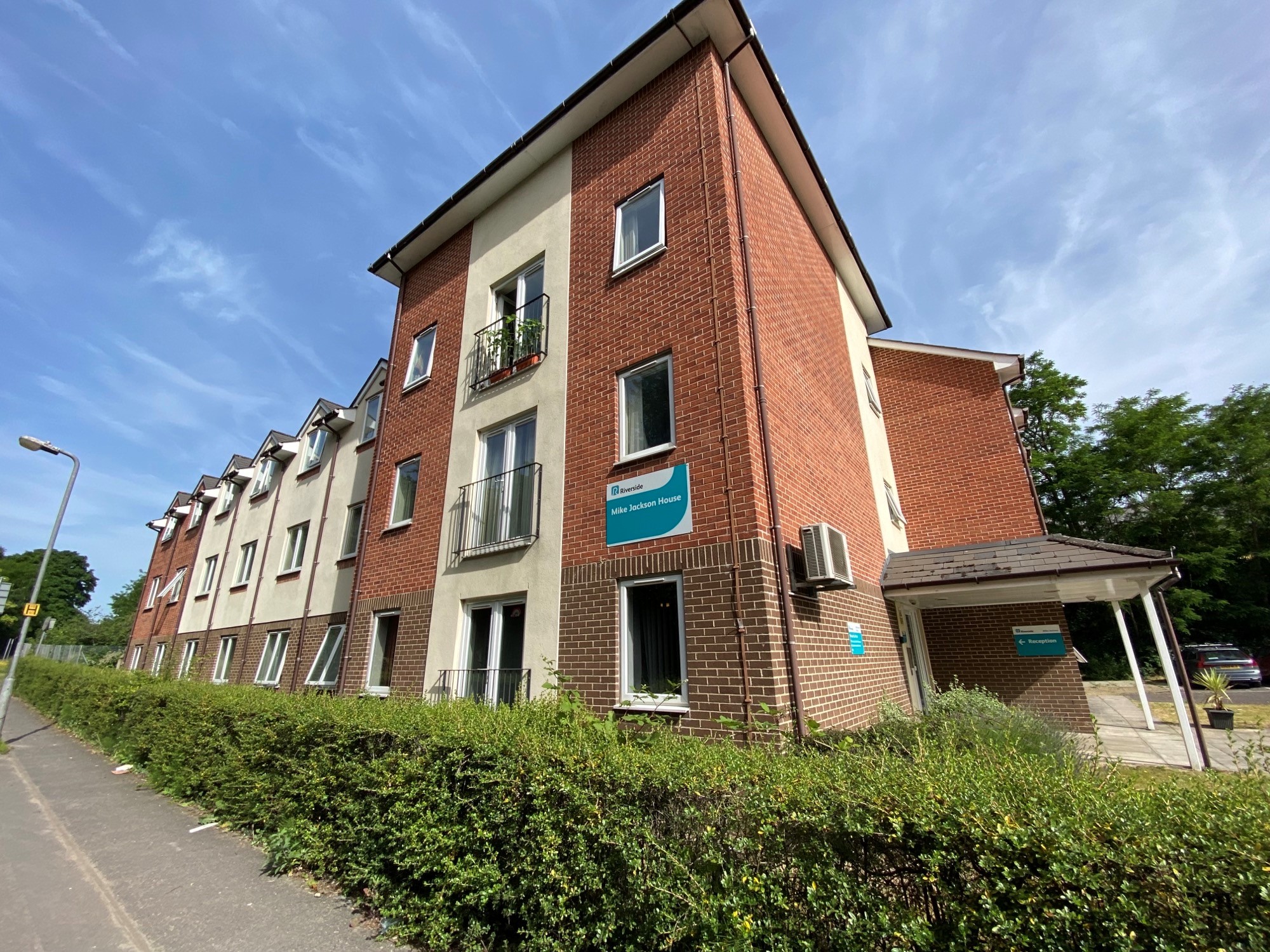 Exterior of a modern three‑storey red‑brick and cream‑rendered residential building with white‑framed windows and small balconies. A sign on the wall reads “Mike Jackson House,” with a covered entrance marked “Reception” to the right. A hedge lines the pavement in the foreground, with trees and a clear blue sky behind the building.
