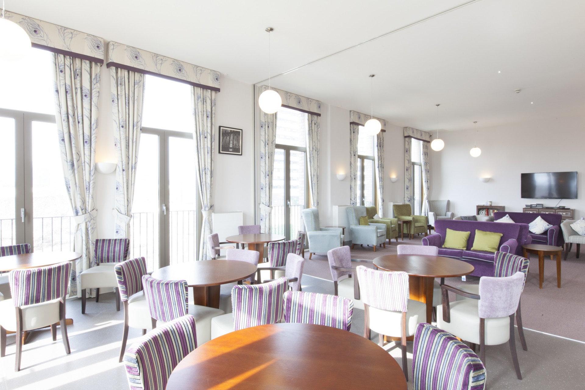Bright communal lounge with round tables and upholstered chairs arranged near tall windows with patterned curtains. A seating area with armchairs and a purple sofa sits further back, with a wall‑mounted television and soft pendant lighting creating a spacious shared environment.