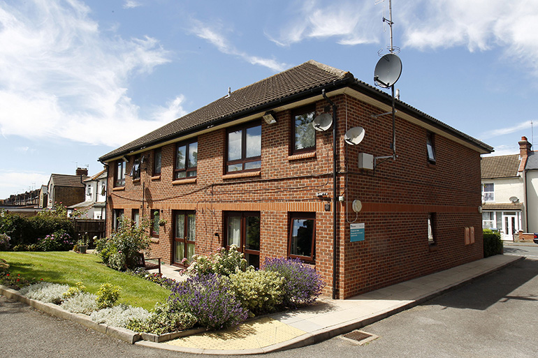 Exterior of a two‑storey red‑brick residential building on a corner plot, with white‑framed windows and a tiled pitched roof. Satellite dishes are mounted on the walls, and a landscaped garden with flowering shrubs and low plants borders the pavement, with neighbouring houses visible along the street under a bright sky.