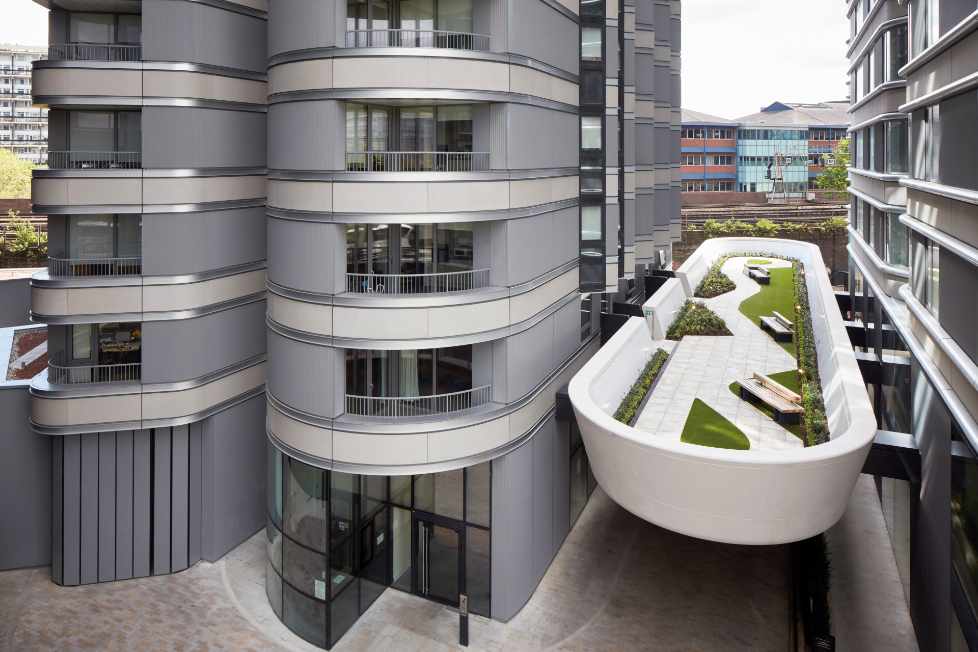 Modern multi‑storey building with curved grey cladding and balconies, viewed from above. A raised landscaped terrace with seating, paved walkways and green planting runs between the buildings, with rail lines and additional buildings visible in the background.