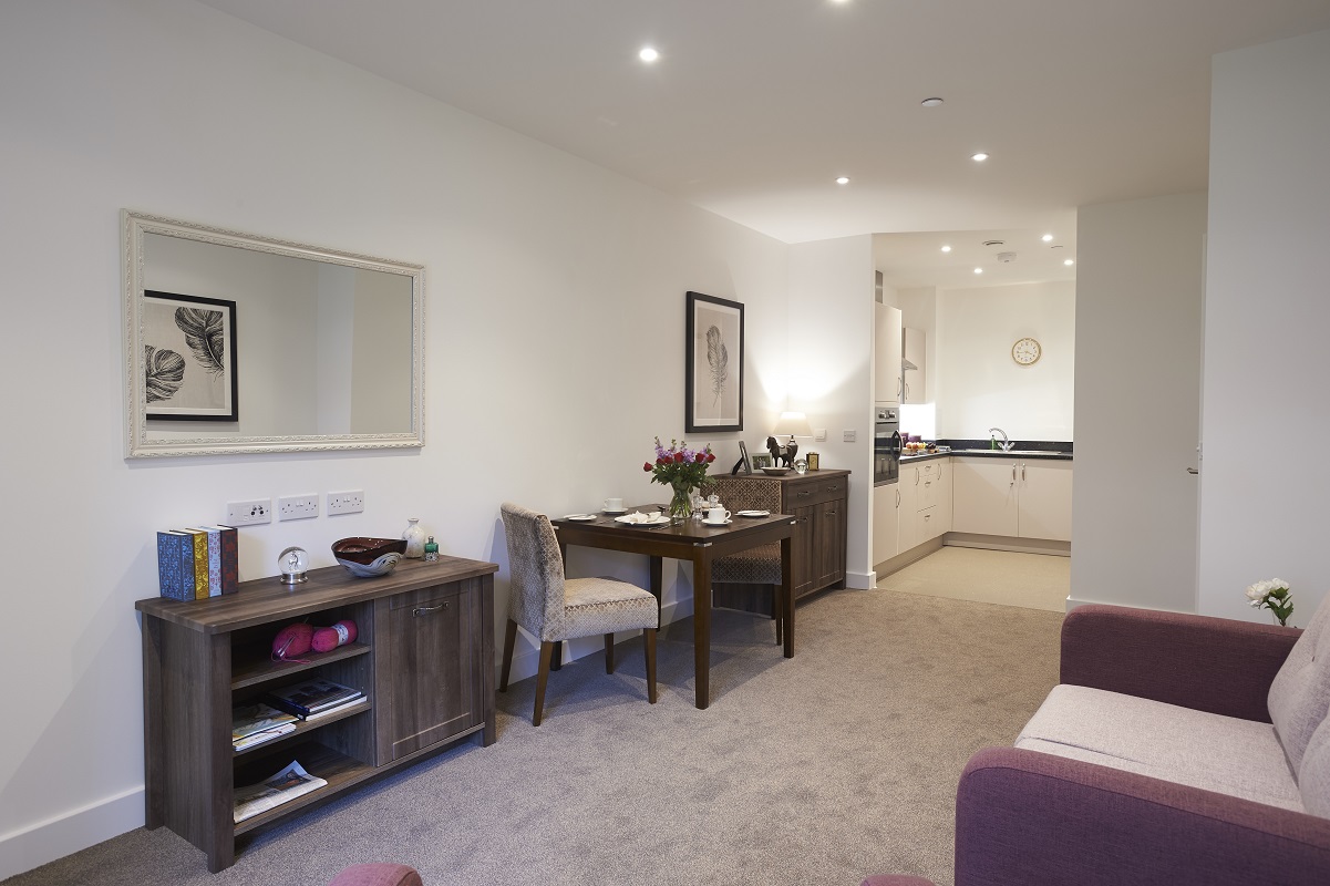 Open‑plan apartment interior with a small dining table set for two, a sideboard and a large wall mirror. A compact fitted kitchen with white cabinets sits at the back, with neutral carpeting and modern ceiling lighting throughout.
