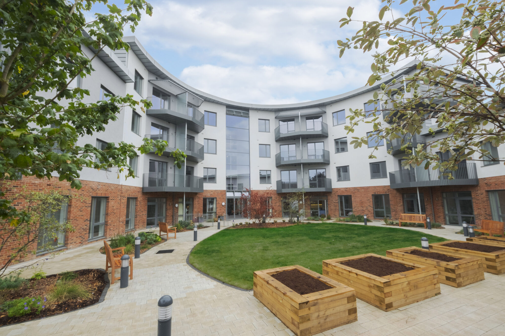 Communal courtyard of a modern multi‑storey residential building with white upper walls and red‑brick ground floor. Curved façades with glass‑fronted balconies overlook a circular lawn, with paved paths, benches and raised wooden planting beds in the foreground, framed by leafy trees under a partly cloudy sky.