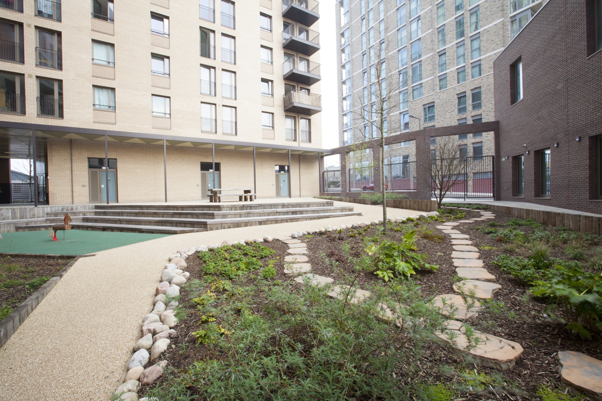 Landscaped courtyard between modern multi‑storey residential buildings, featuring a curved pathway, planted beds and a small tree. Steps lead up to a raised seating area with benches, and a play feature sits to the left, with surrounding apartment buildings and balconies visible.
