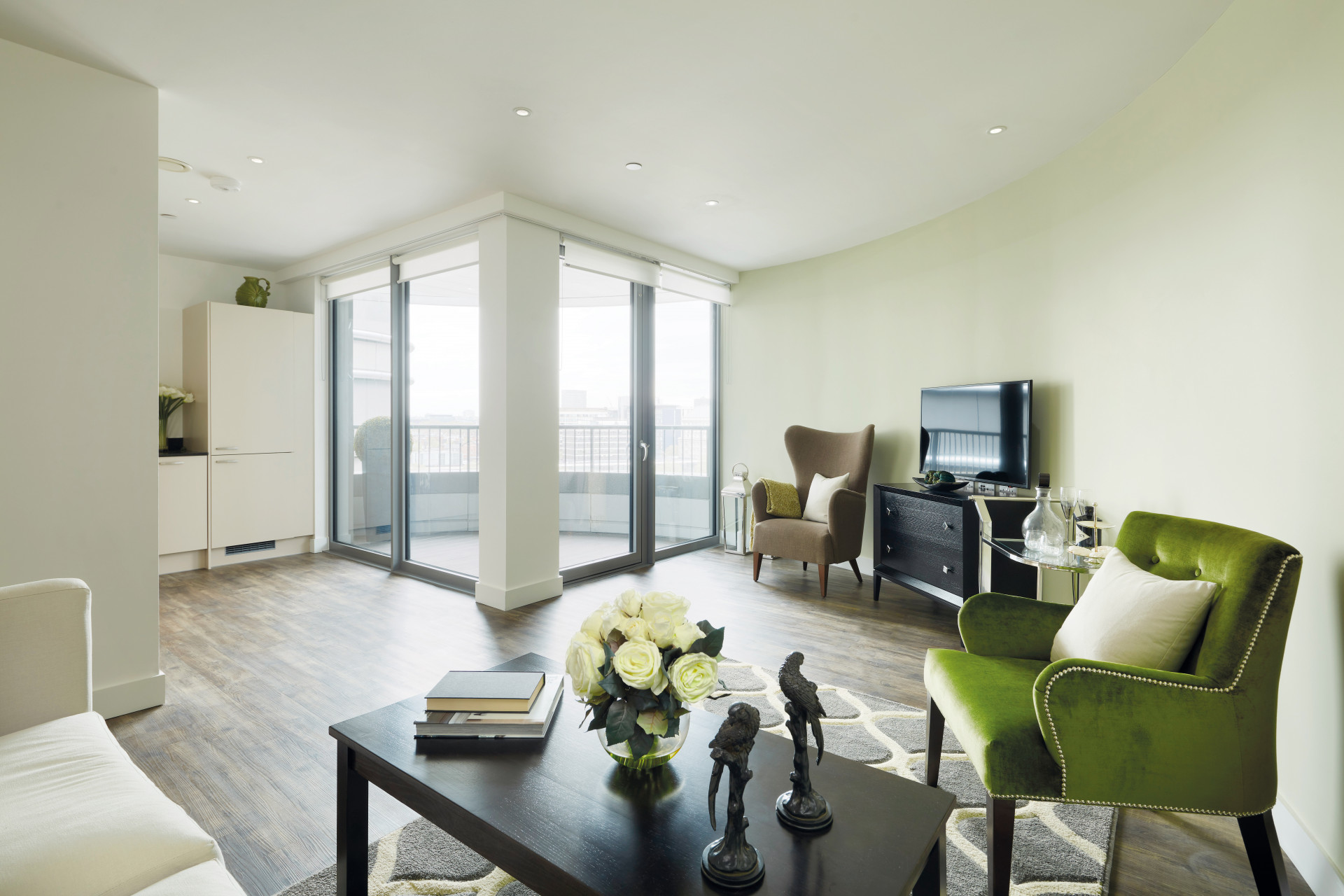 Modern open‑plan living area with wood‑effect flooring and neutral décor, featuring armchairs, a coffee table with decorative items and a TV unit. Floor‑to‑ceiling glass doors open onto a balcony, allowing natural light into the space.