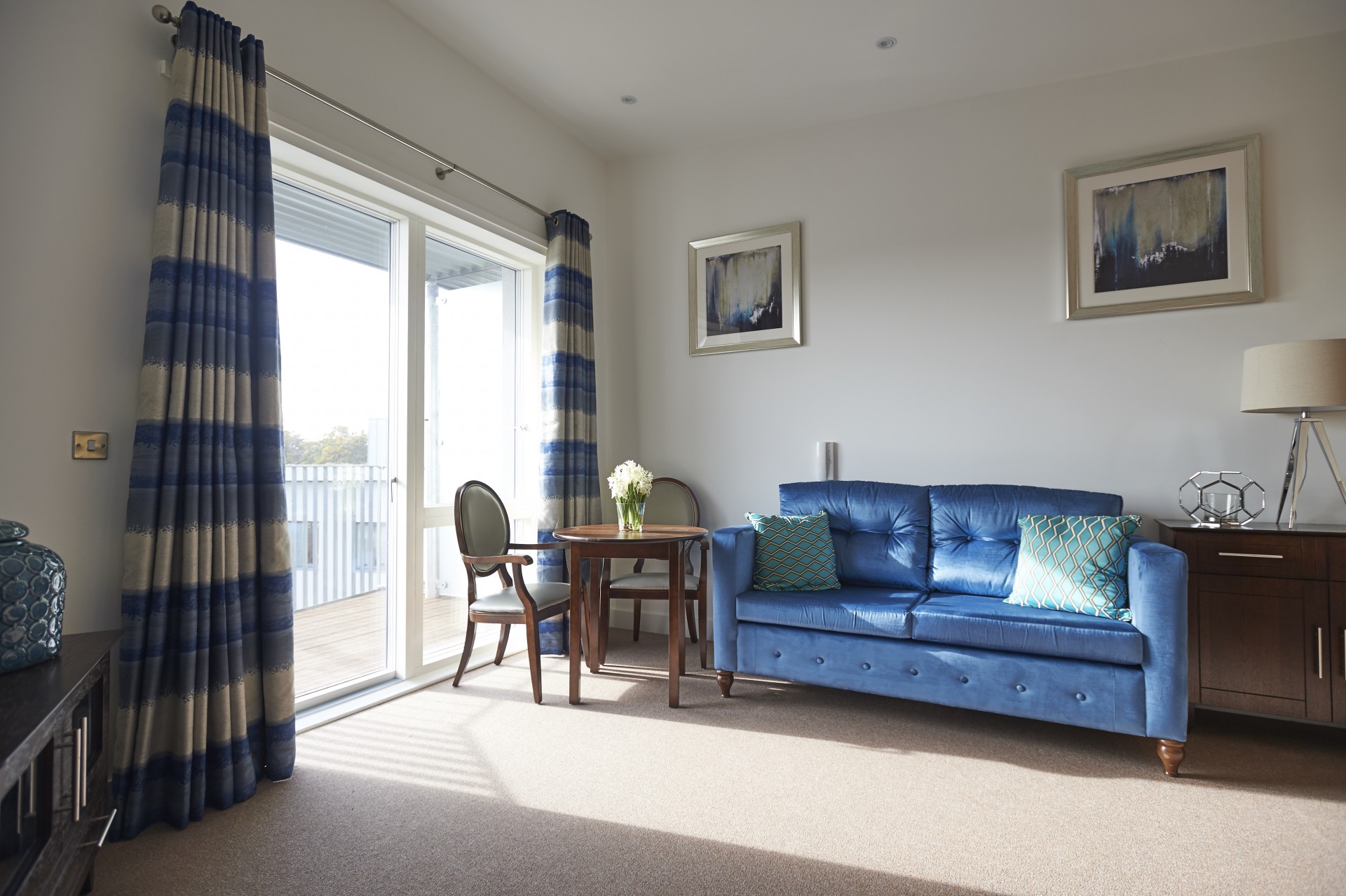 Living room with a blue upholstered sofa and patterned cushions, beside a small round dining table and two chairs. Large glass doors with striped curtains open onto a balcony, letting natural light into the carpeted space, with framed artwork and a side table along the wall.
