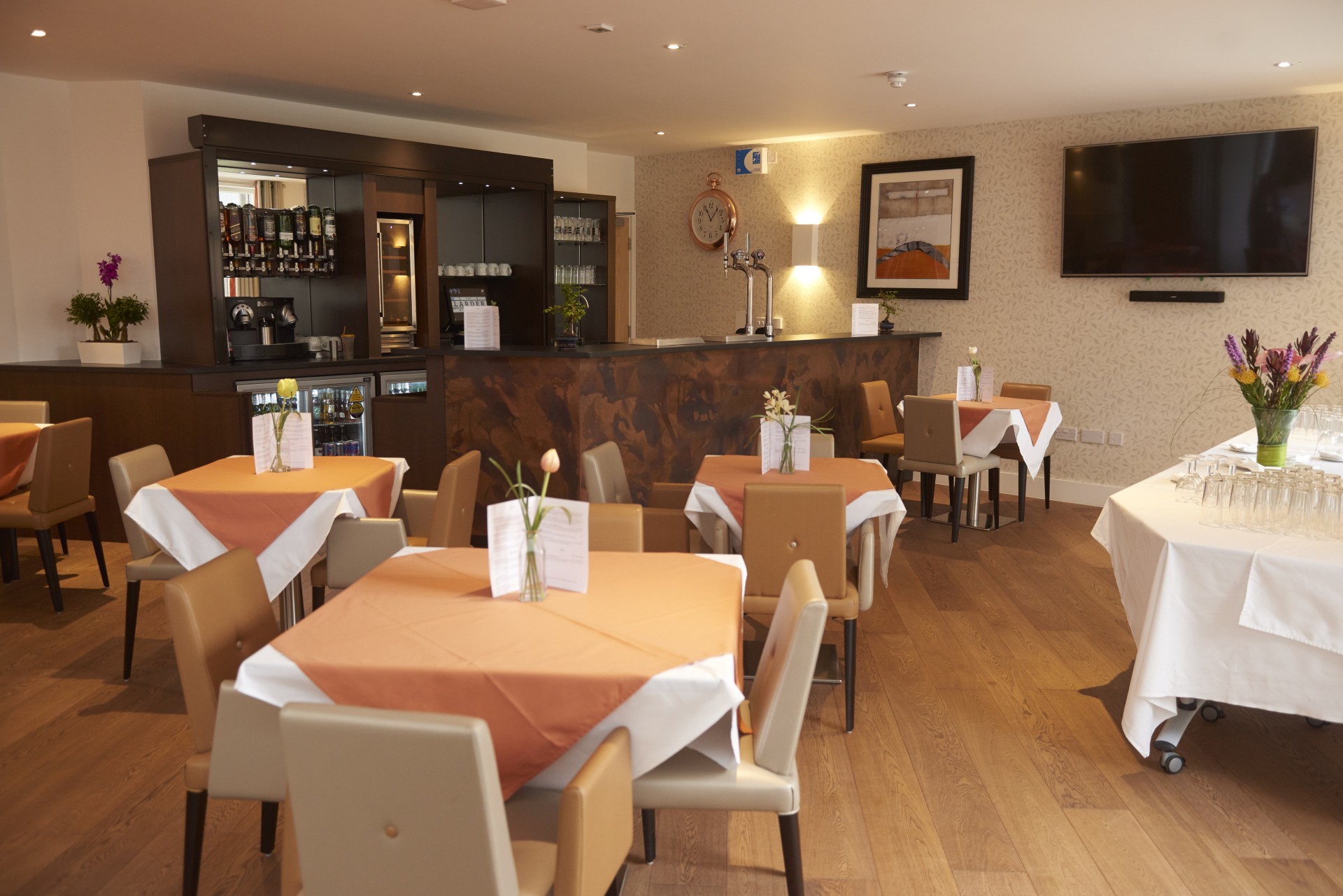 Communal dining room with square tables set with orange and white table cloths and small vases of flowers. Upholstered chairs surround the tables, with a dark wood bar area and drinks display at the back, a wall‑mounted television, framed artwork and warm lighting throughout.