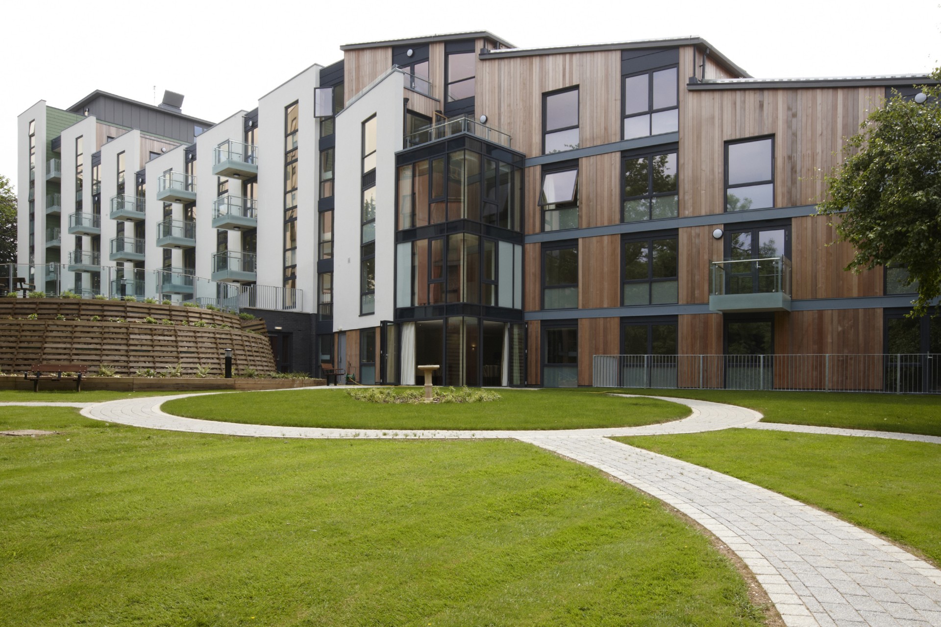 Communal garden courtyard in front of a modern residential building with white upper walls and timber‑clad sections. Curved balconies overlook a central circular lawn, with paved paths, benches and raised wooden planting beds in the foreground, framed by trees under a cloudy sky.